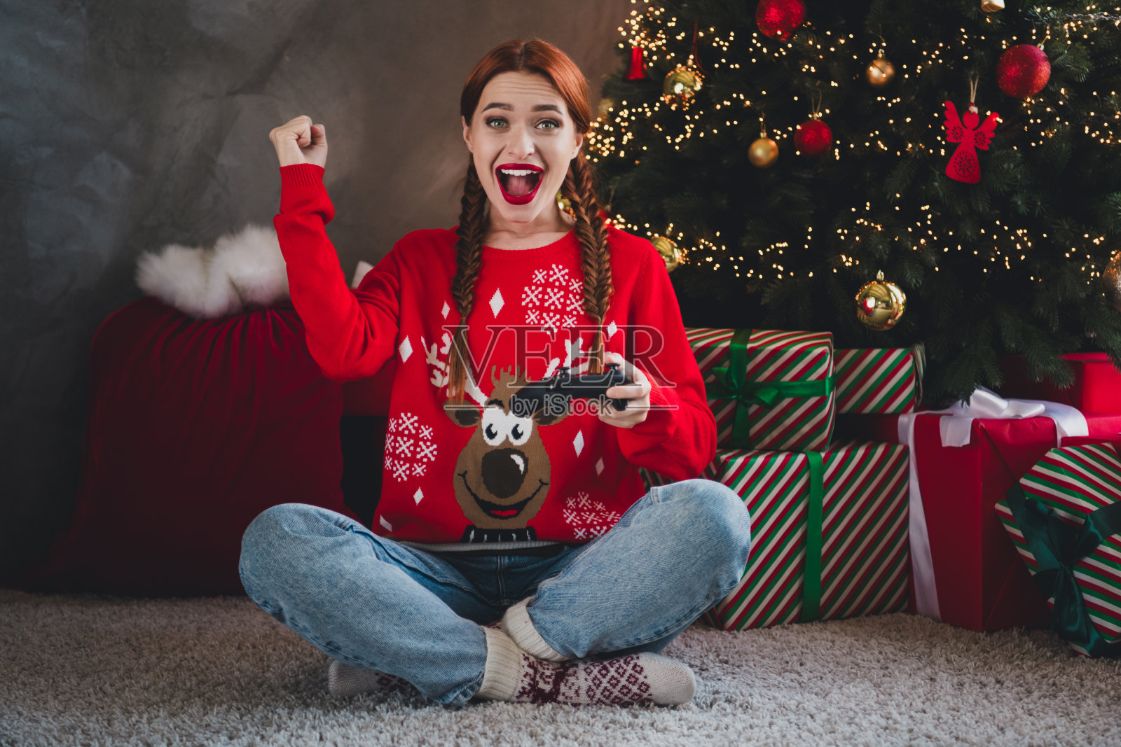 年轻的红发女性穿着圣诞服装，在室内房间的传统氛围装饰中，兴奋地玩电子游戏并获胜的全身照片照片摄影图片