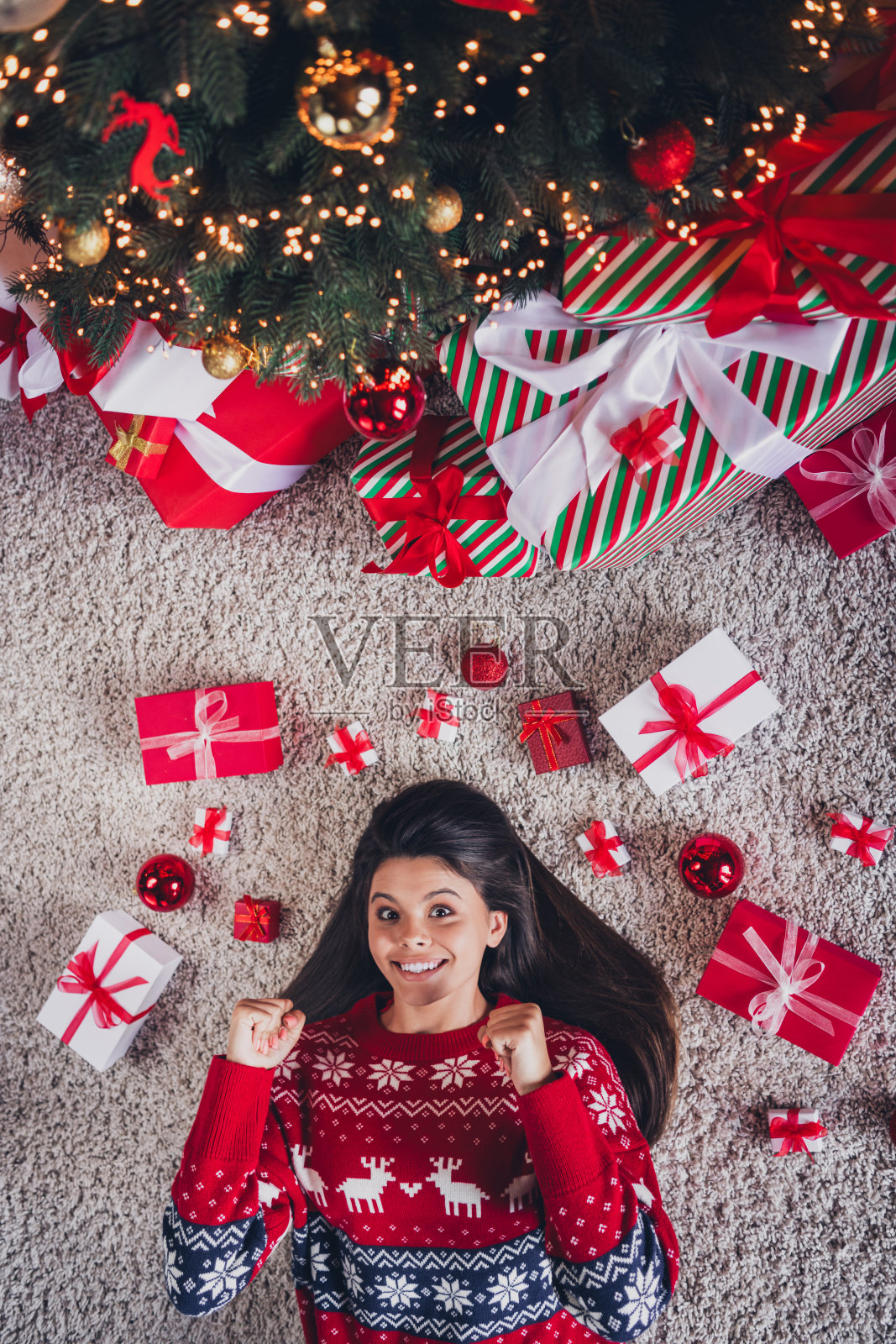 高角度拍摄可爱女孩穿着新红色印花衣服，躺在地毯上享受室内圣诞节装饰的照片。照片摄影图片