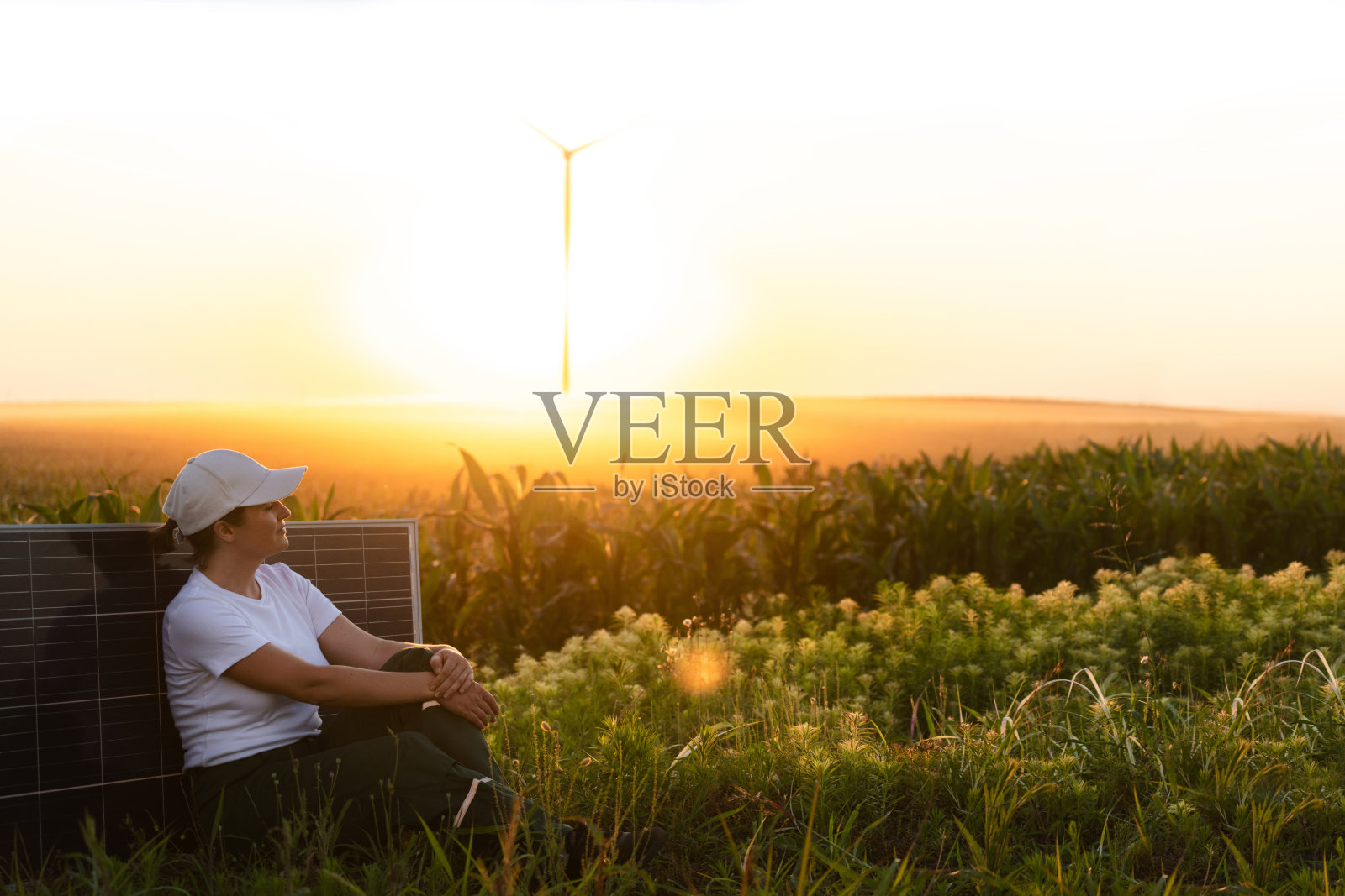 穿着白色帽子的女人坐在日落时分的太阳能电池板旁，背景是风力涡轮机。照片摄影图片