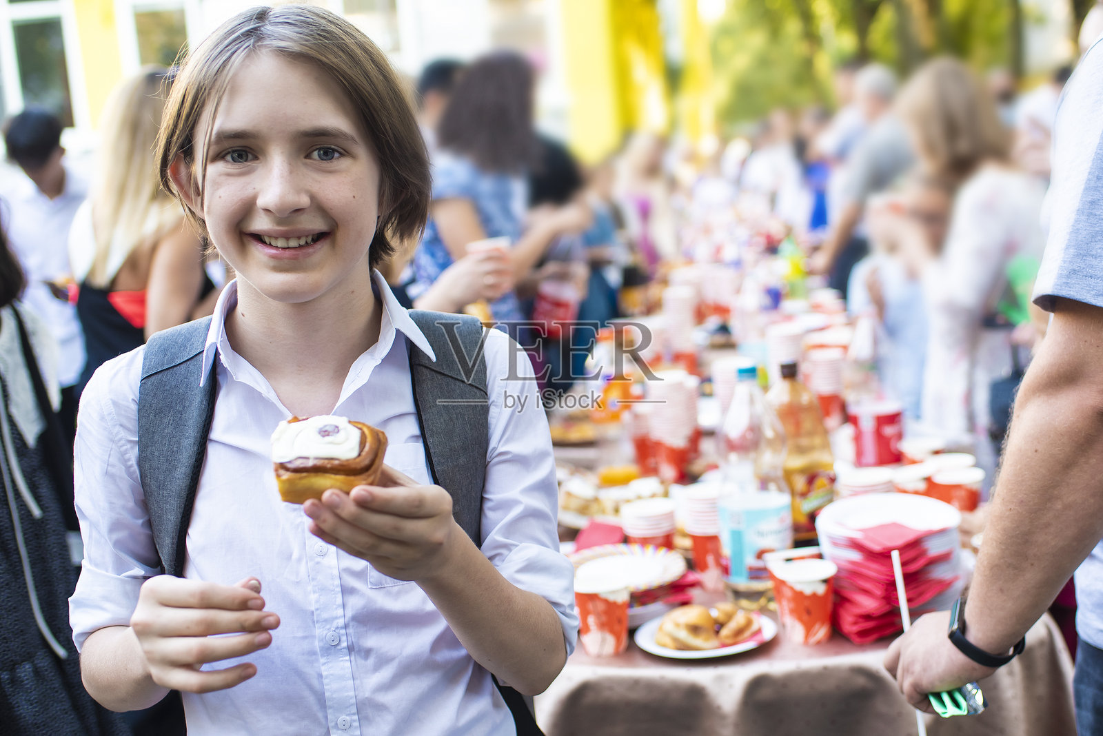 一个穿着校服、手里拿着书包的男孩在学校的茶会上，站在节日餐桌前，手里拿着一个包子。照片摄影图片