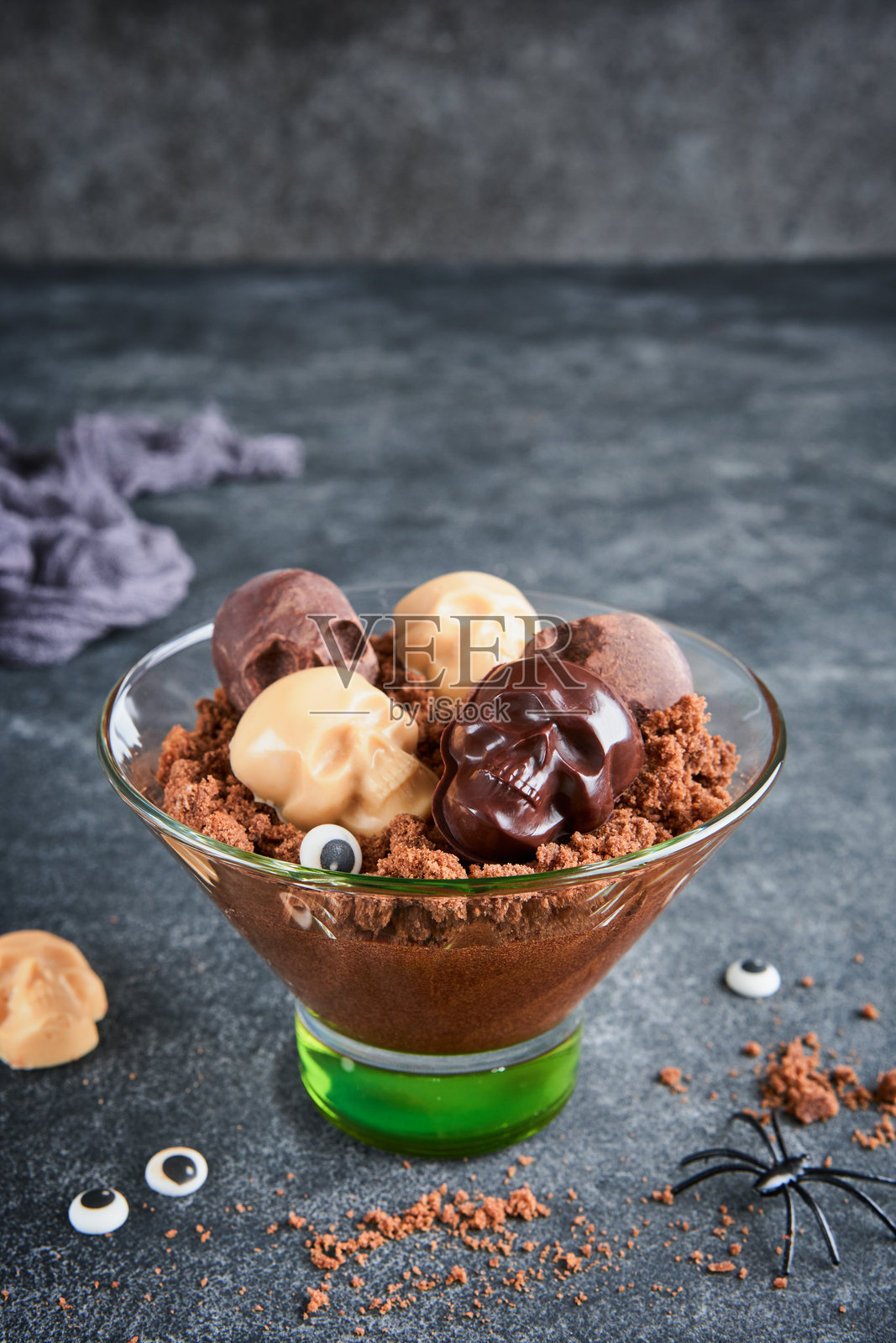 万圣节派对巧克力布丁或意式奶冻，配有布朗尼碎屑和巧克力骷髅，盛放在透明玻璃杯中，背景为深灰色。万圣节派对创意。照片摄影图片