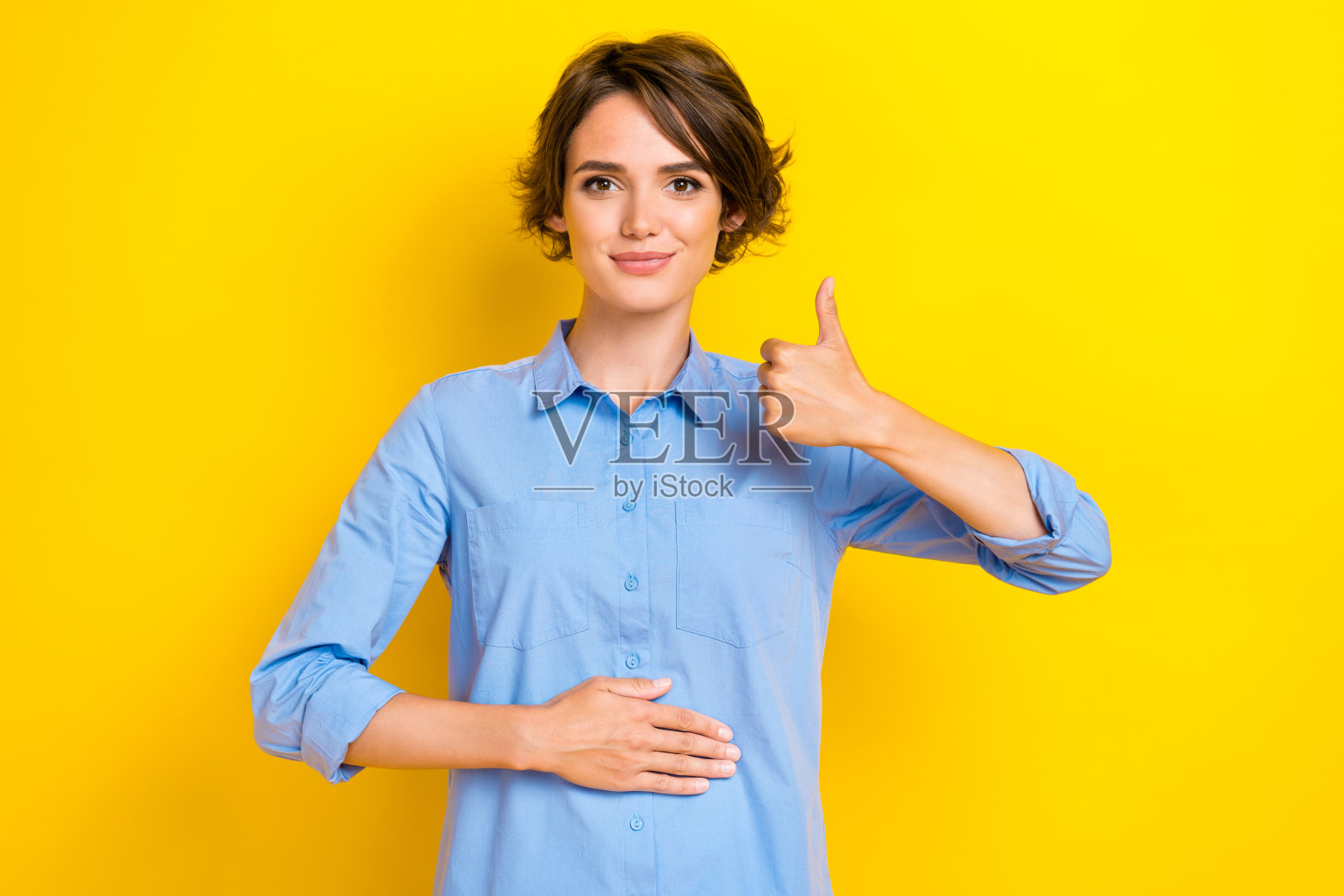 自信积极的女士穿着蓝色衬衫，双手放在腹部，竖起大拇指，背景为黄色。照片摄影图片