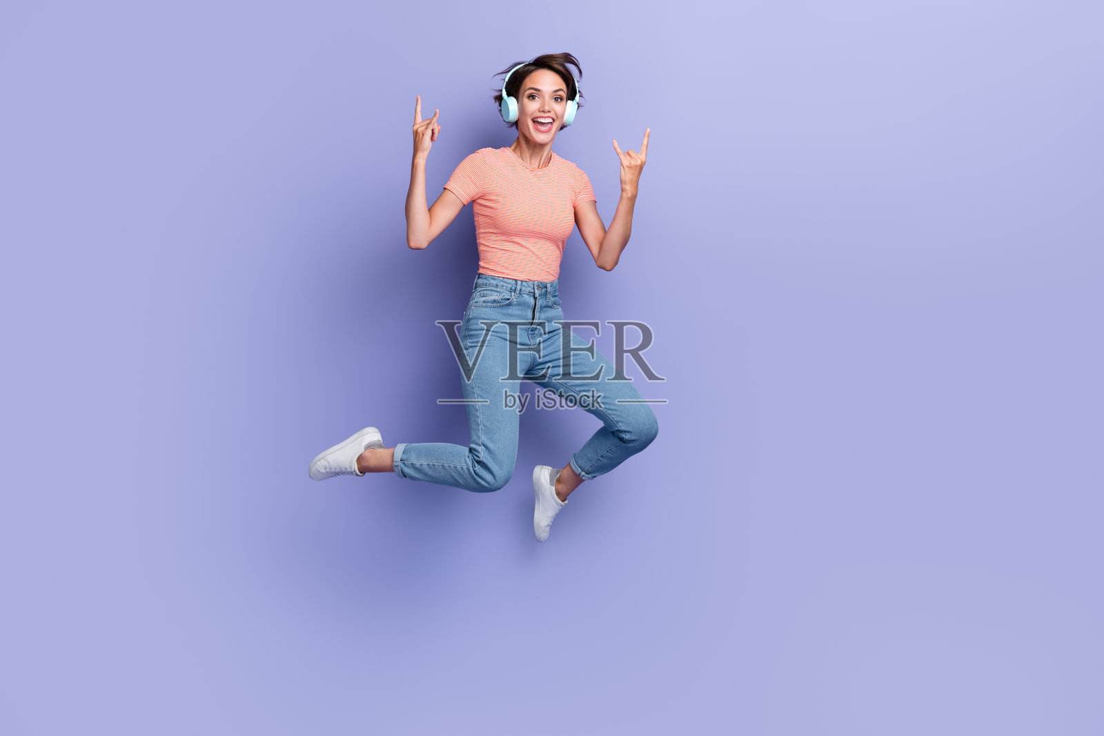 全身照，酷炫的女孩穿着橙色T恤，戴着耳机，跳得很高，做出摇滚手势，背景为紫色。照片摄影图片