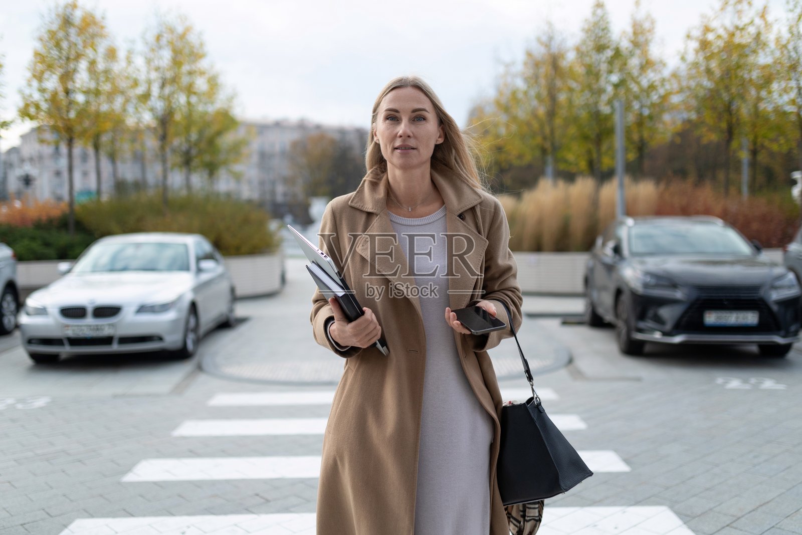 一位女商人在商务中心附近走着，手里拿着笔记本电脑和手机，显得自信且准备迎接新的一天。照片摄影图片