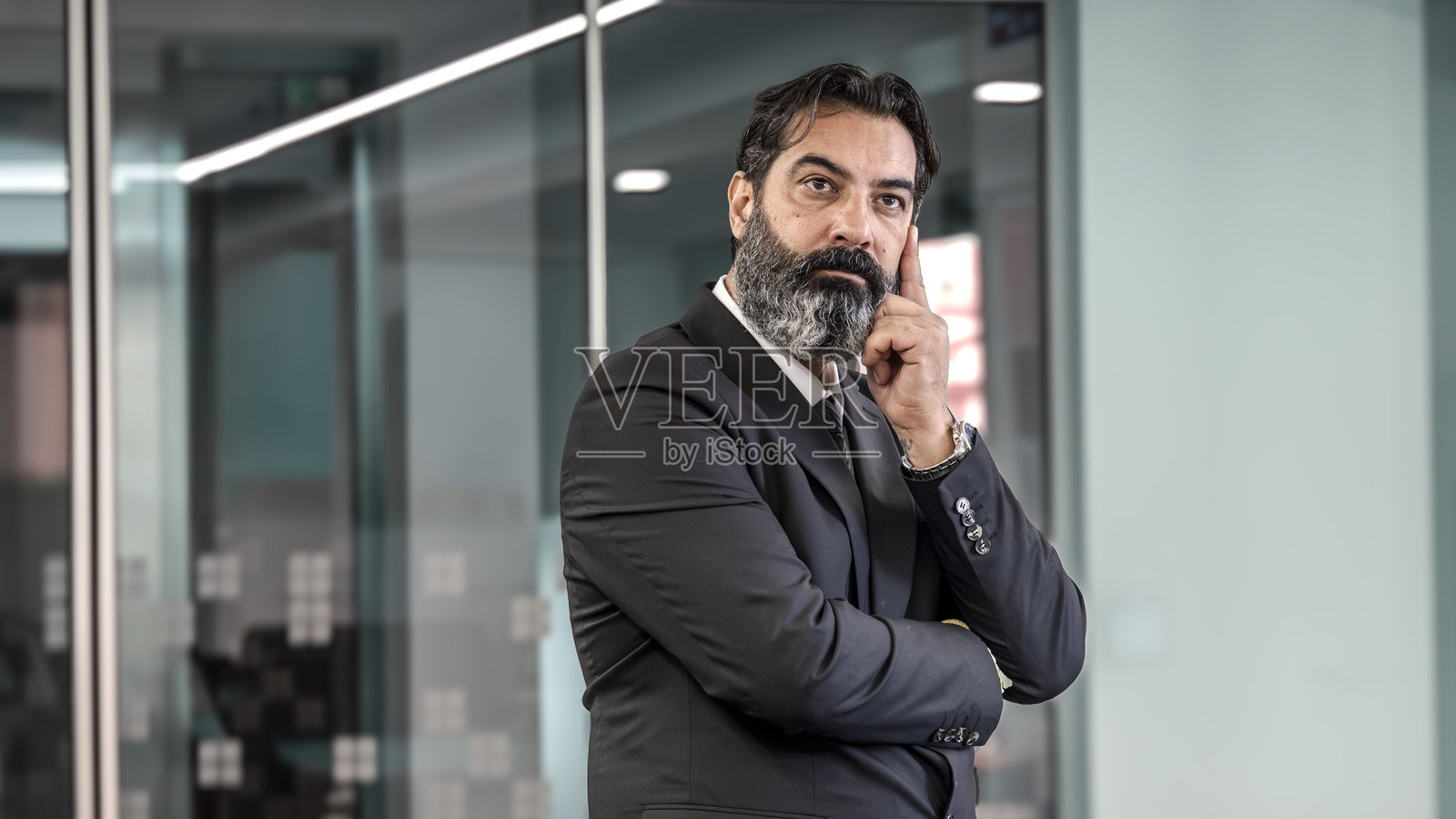 一位中年、深色头发、留着胡子的商人在一个空旷的办公室环境中，背景模糊，特写镜头中，他用手托着脸颊，面部表情显示他在思考。照片摄影图片