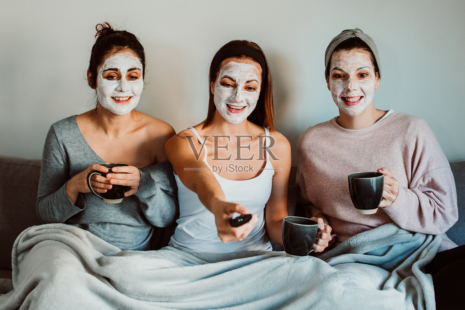 快乐的女性朋友们在家靠墙坐着，戴着面膜看电视照片摄影图片