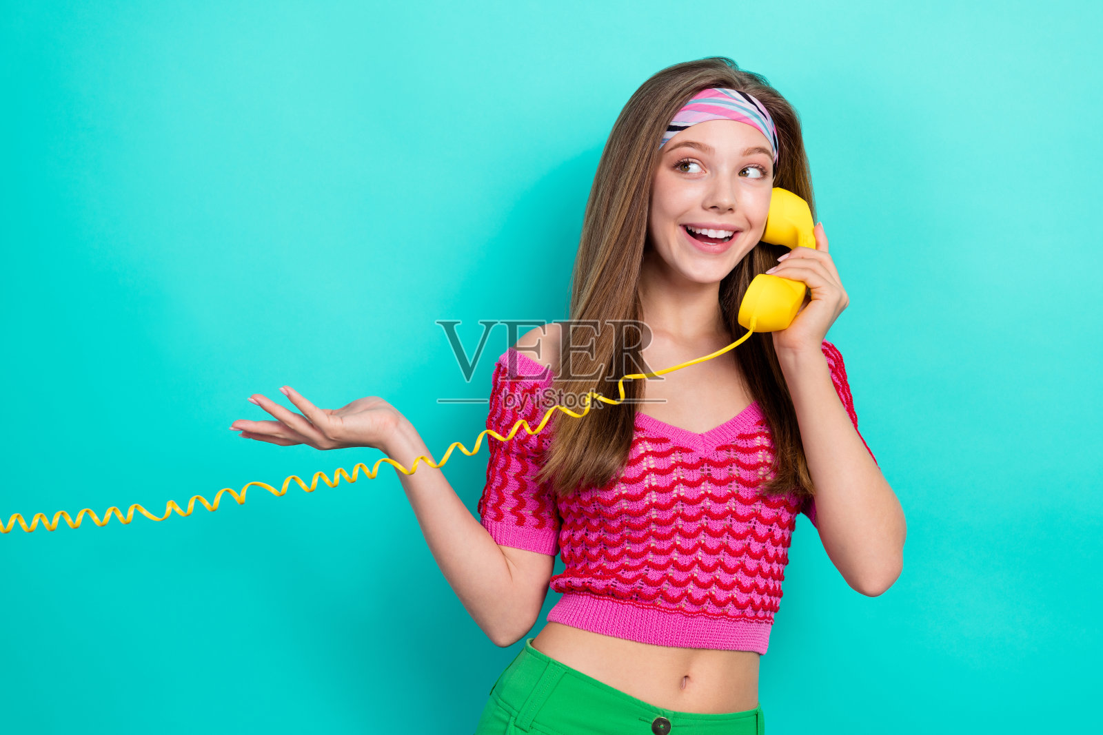 一位积极、好奇的金发女孩穿着粉色针织上衣，正在蓝绿色背景下使用座机电话交谈。照片摄影图片