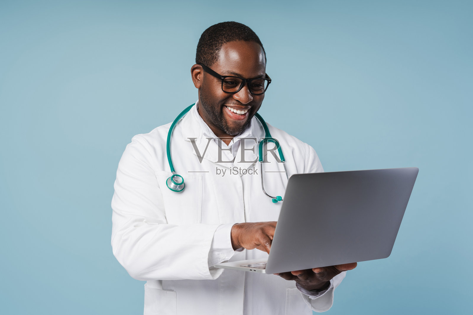 微笑的非裔美国医生戴着眼镜，在蓝色背景的工作室里用笔记本电脑打字。照片摄影图片
