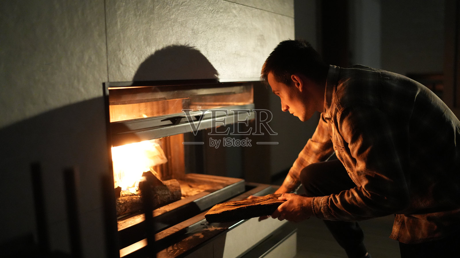 一个男人小心翼翼地将一根木柴放入发光的壁炉中，温暖和光线充满了寒冷夜晚的房间，营造出温馨舒适的氛围。照片摄影图片