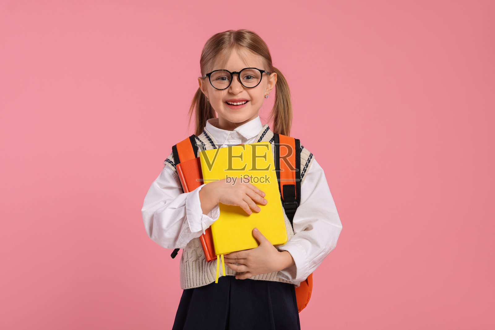 戴着眼镜的快乐女学生，背着书包和书本，背景为粉色。照片摄影图片