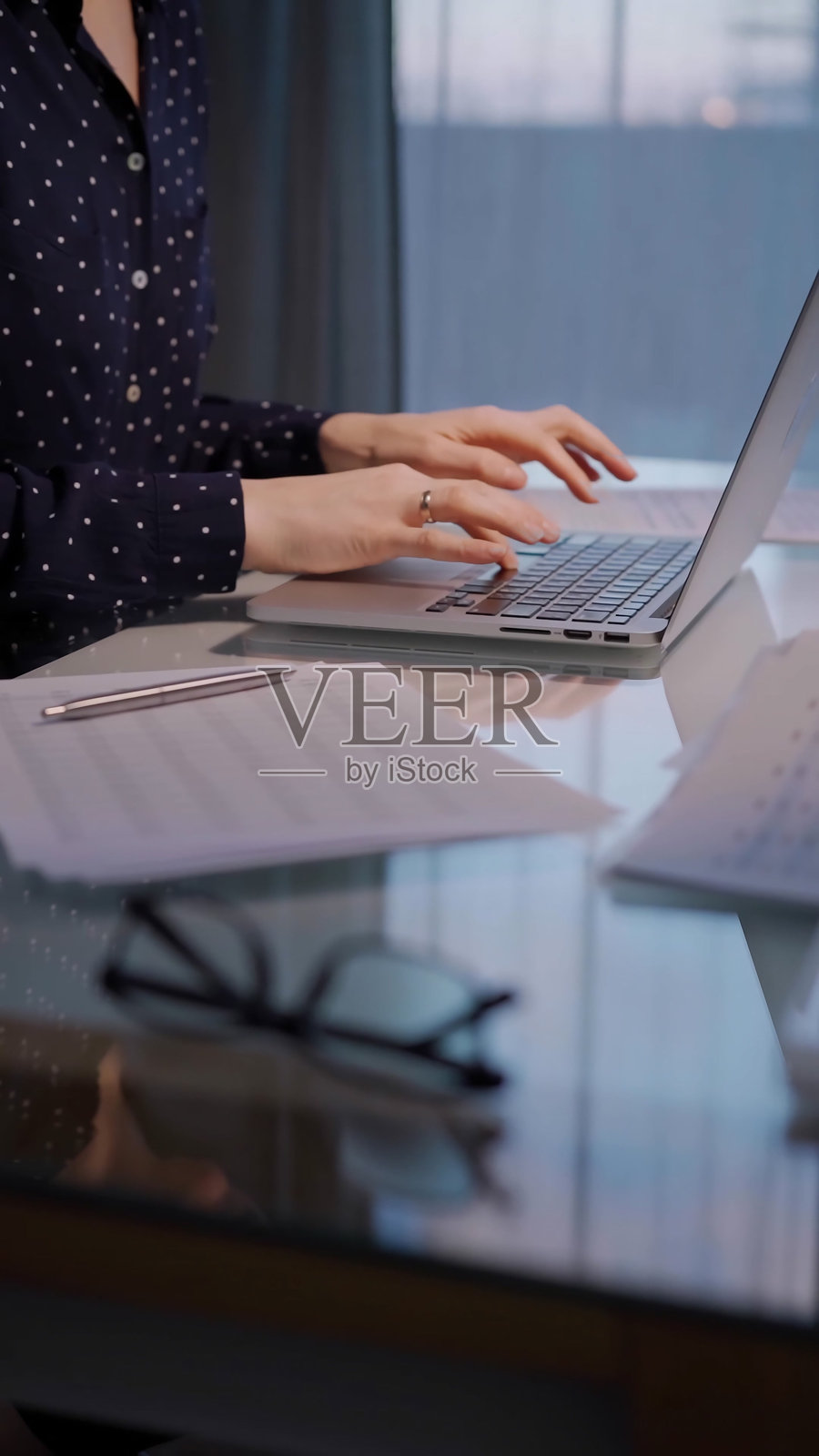 穿着蓝色波点裙的女商人在办公室的玻璃桌旁坐着，傍晚时分专注地在笔记本电脑上打字，特写镜头。照片摄影图片