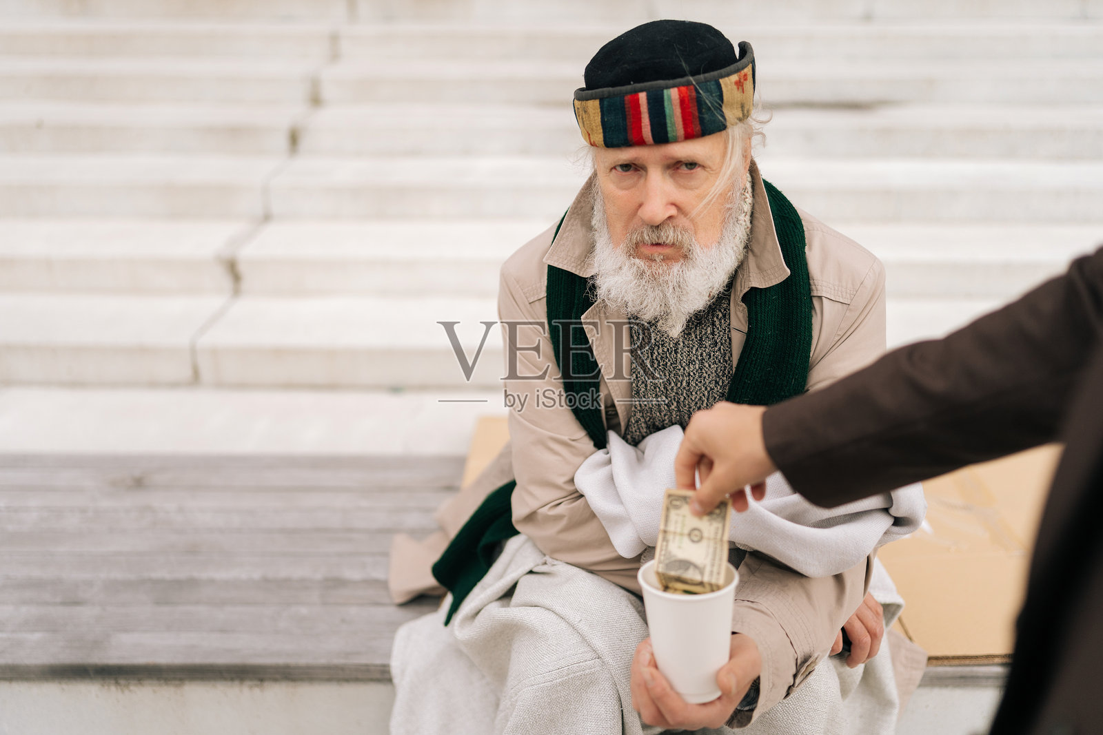 一位悲伤的老年无家可归者坐在城市台阶上，从穿着西装的商人手中接过一美元纸币，放入白色纸杯中。照片摄影图片