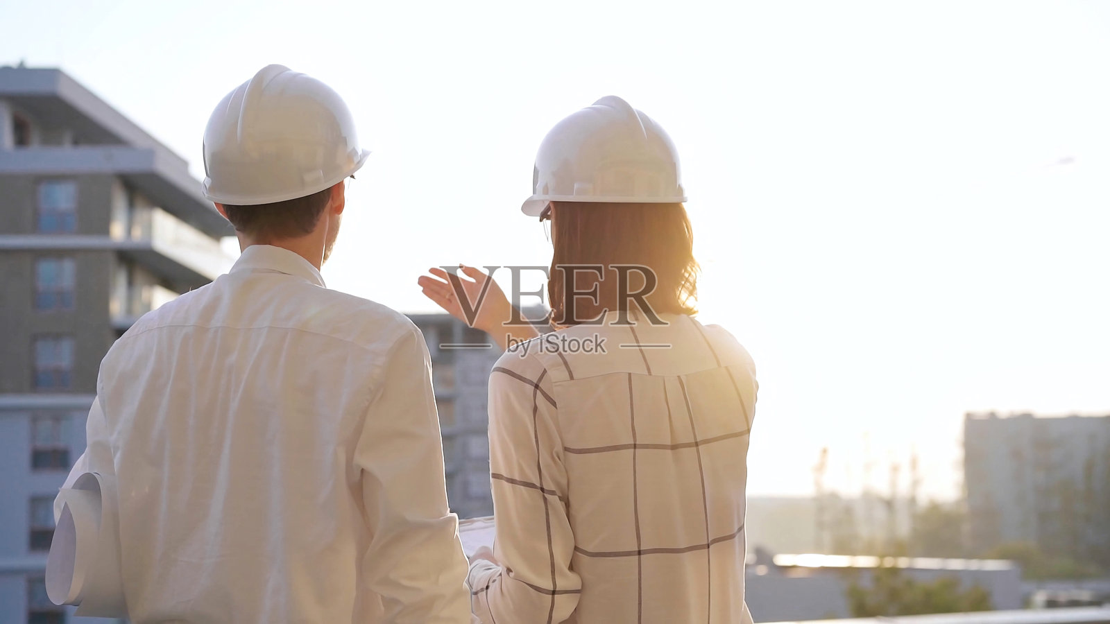 两位建筑师同事，戴着安全头盔和眼镜，在日出或日落时分的建筑工地上查看蓝图，背影。建筑与工程照片摄影图片