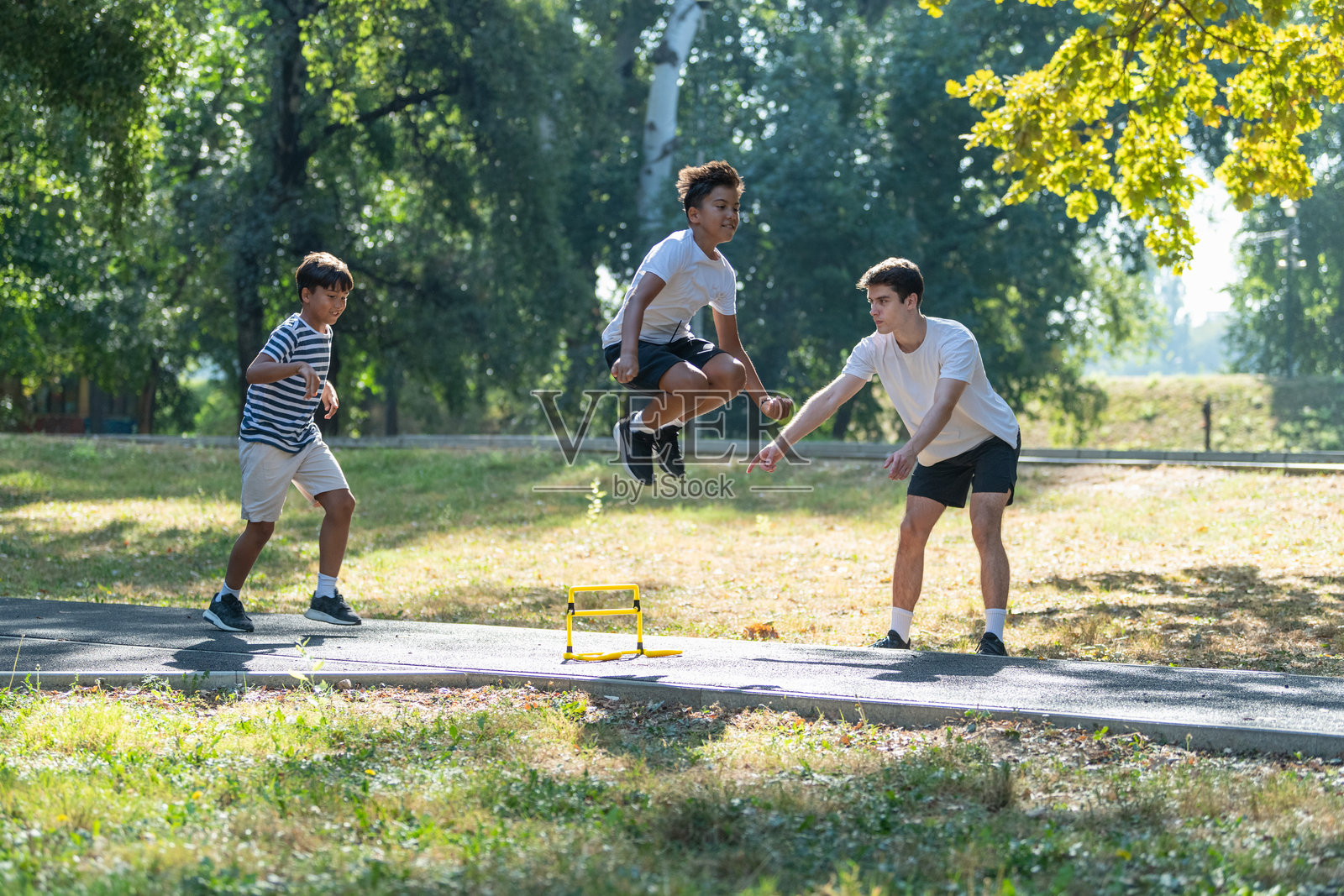 年轻男孩在户外训练课上跳过障碍物，教练在旁指导。该图像展示了旨在提高灵活性和协调性的体育活动，背景为开放的自然环境。照片摄影图片