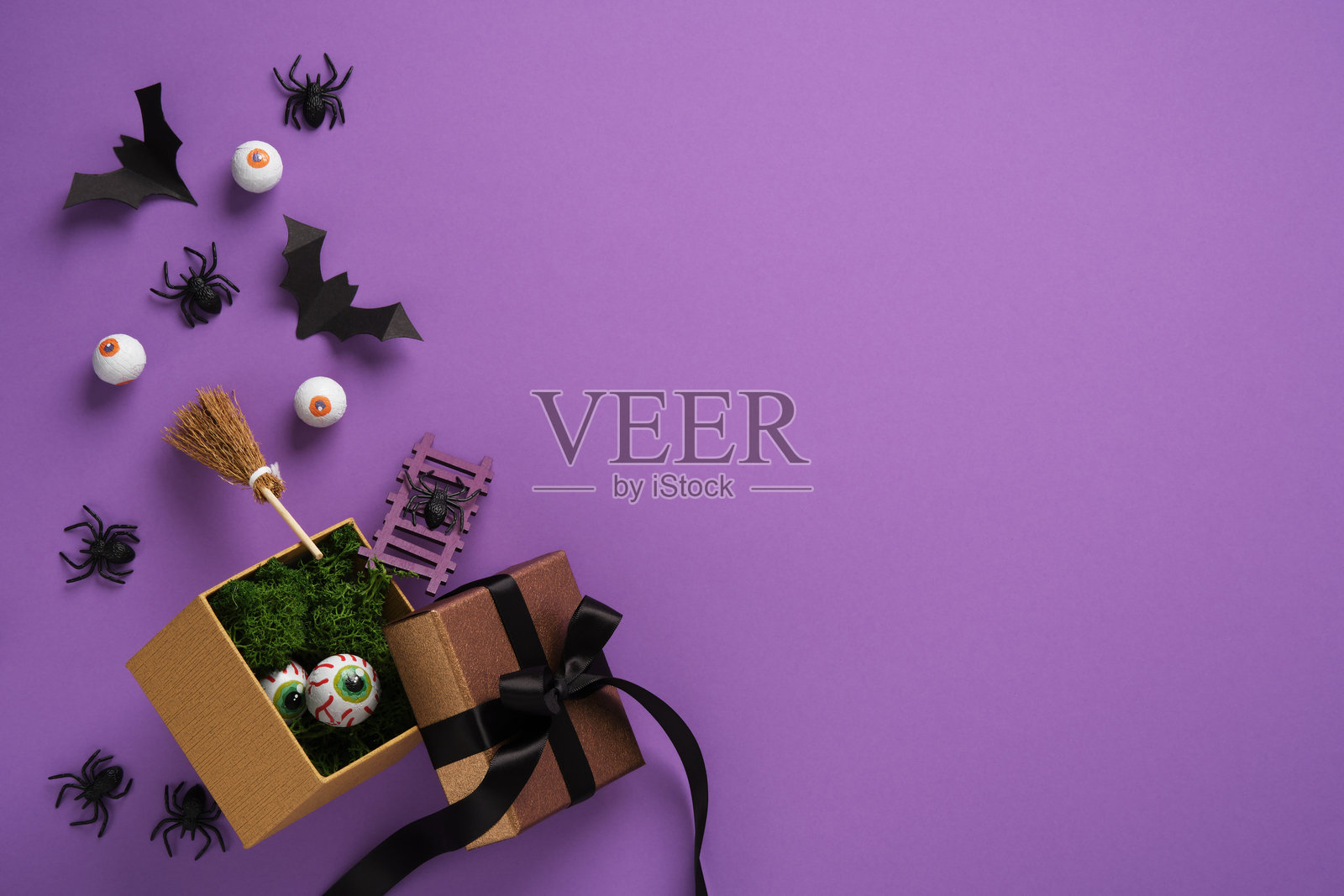 万圣节礼品盒，带有恐怖装饰：假眼球、蝙蝠、蜘蛛和扫帚，背景为紫色。可用于您的文本、公告或邀请。照片摄影图片
