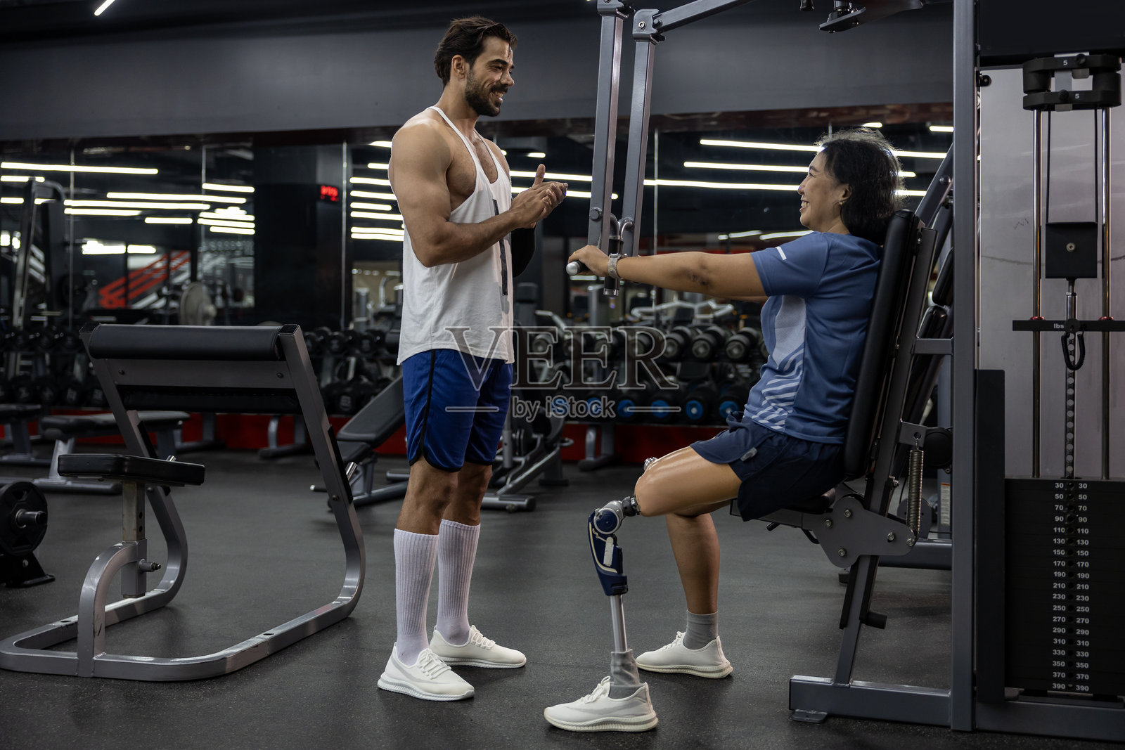 亚洲女性在健身房与专业教练一起使用器械进行力量训练，腿部为假肢，因伤残和康复而进行锻炼。照片摄影图片