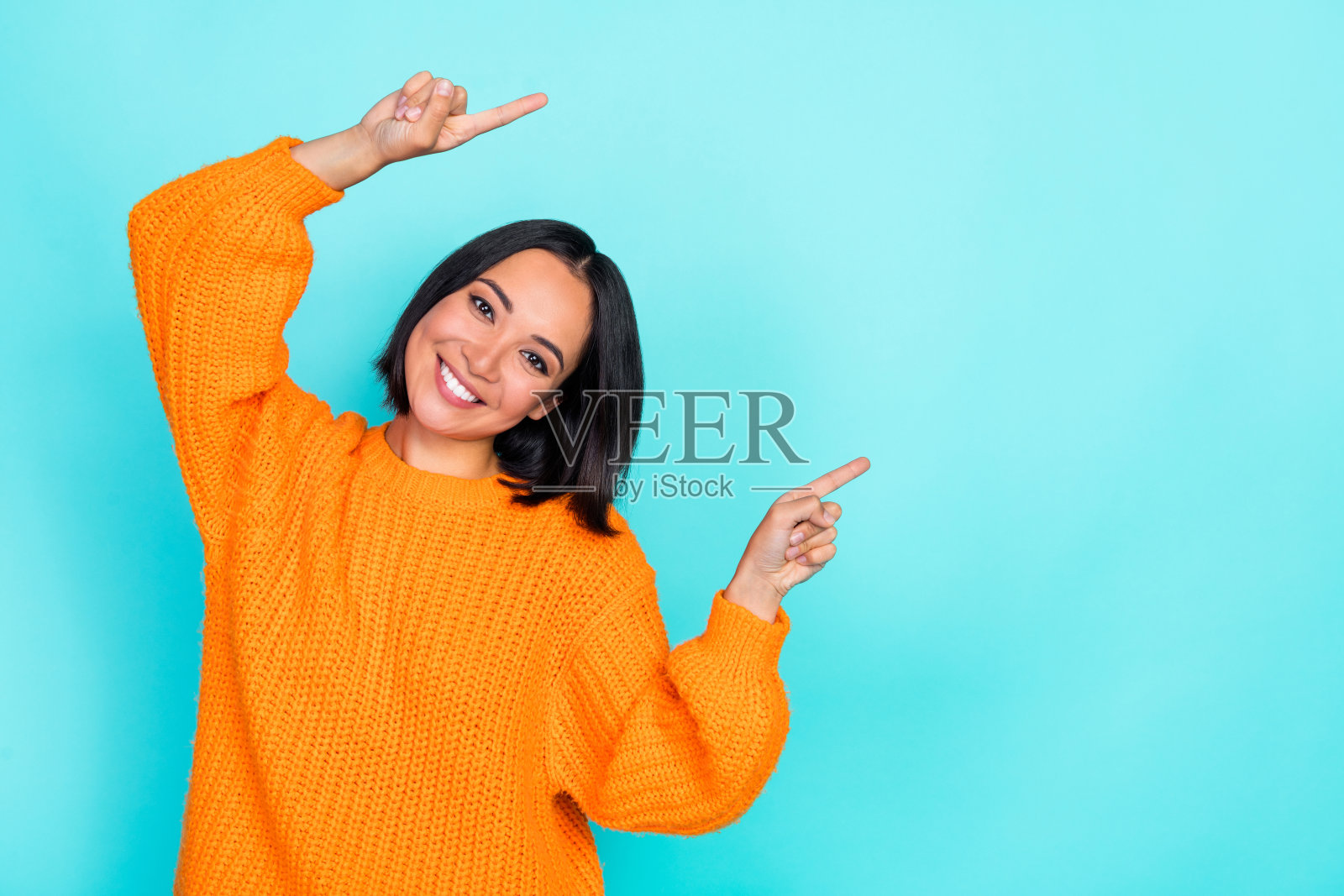 乐观可爱的短发女性肖像，穿着橙色毛衣，指向空白区域，背景为青绿色。照片摄影图片