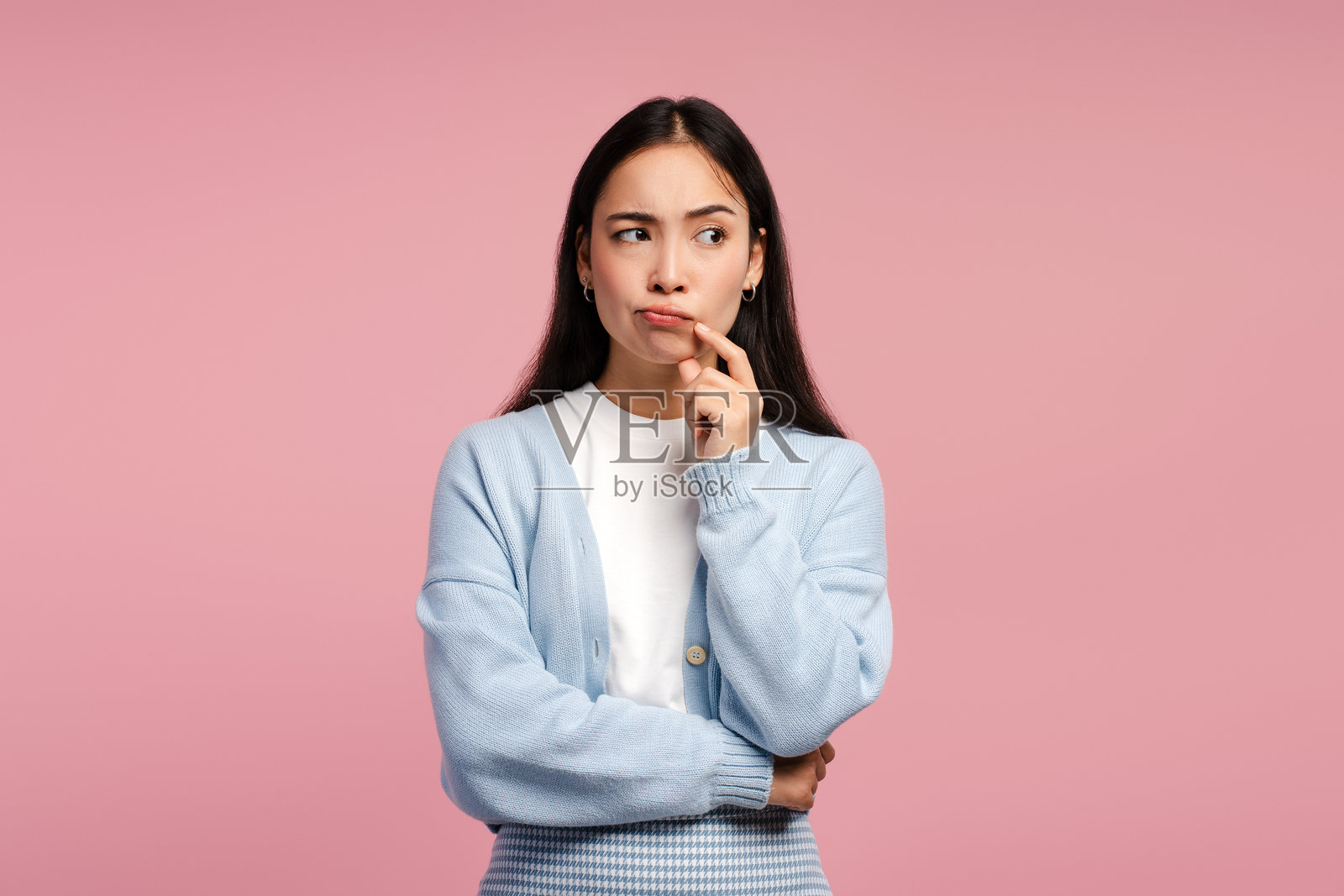 年轻女性深思熟虑，手托下巴，摆姿势于粉色背景，目光投向一旁。照片摄影图片