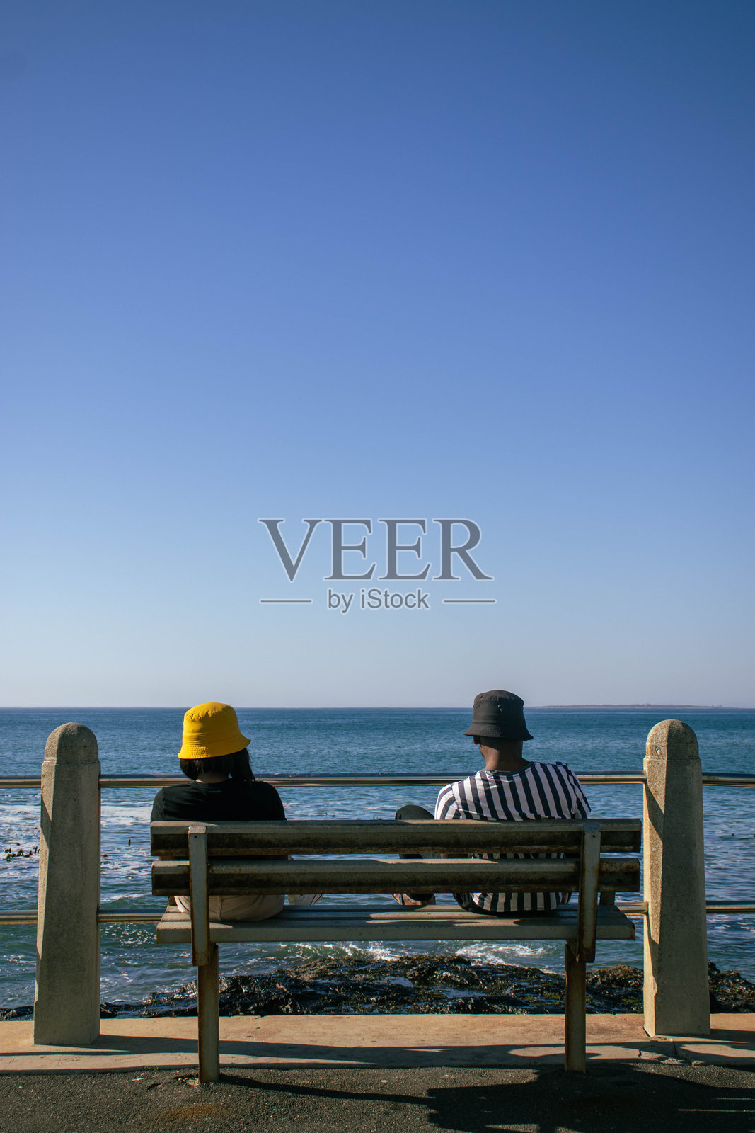 一对年轻情侣坐在长椅上,望着晴朗夏日的海洋。照片摄影图片