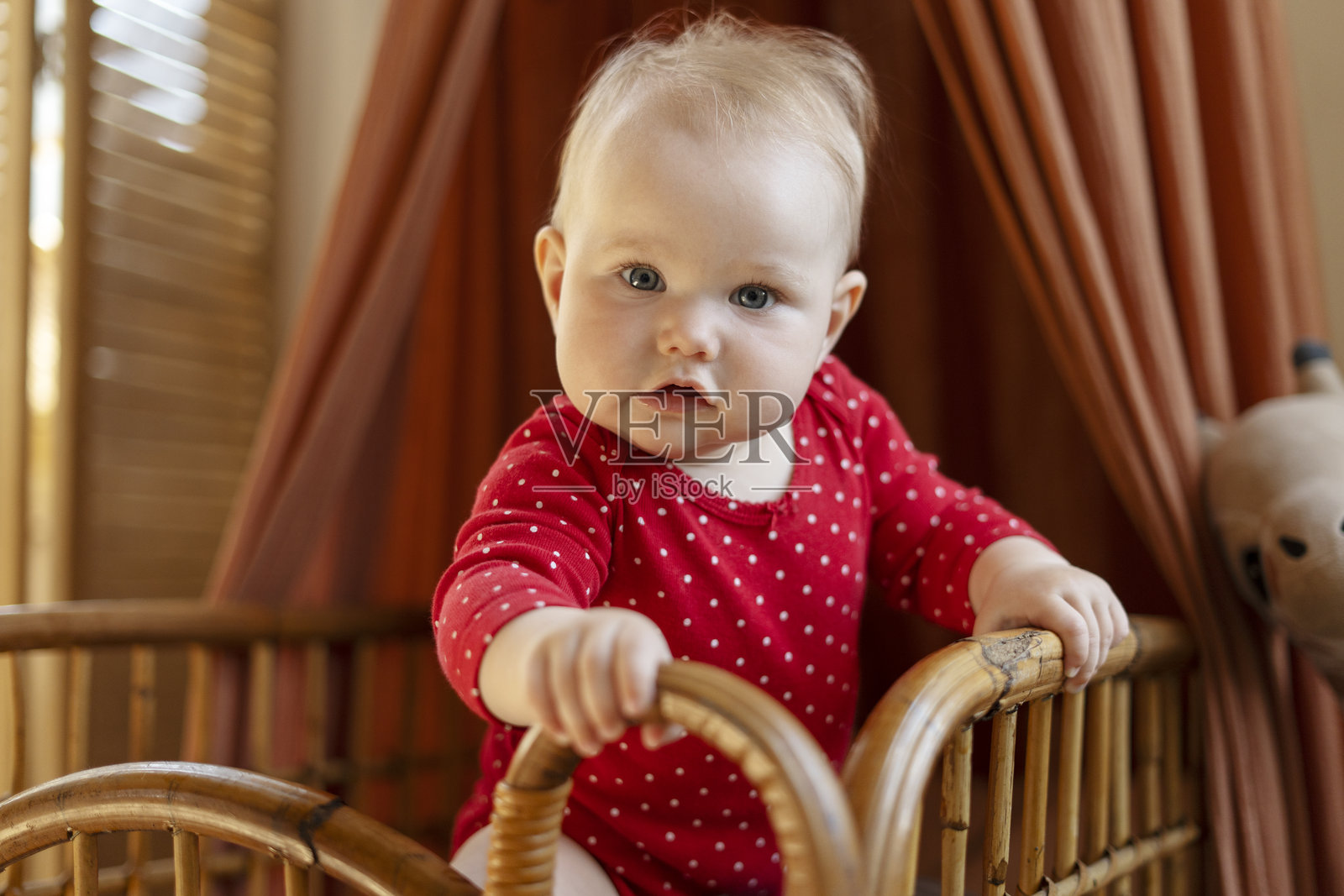 穿着红色连体衣的婴儿女孩站在柳条婴儿床里照片摄影图片