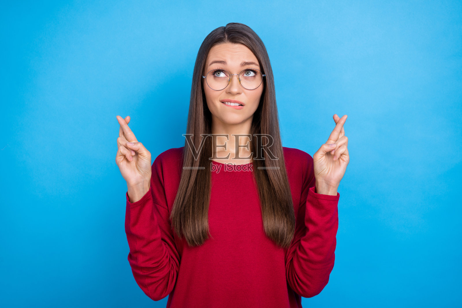 一位年轻女士穿着酒红色毛衣，戴着眼镜，焦虑地仰望，背景为蓝色。照片摄影图片