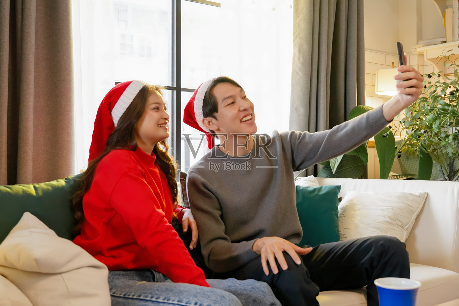 一对亚洲情侣戴着圣诞老人帽，坐在沙发上微笑着一起自拍，捕捉在温馨的客厅中庆祝节日圣诞时的快乐时刻。照片摄影图片