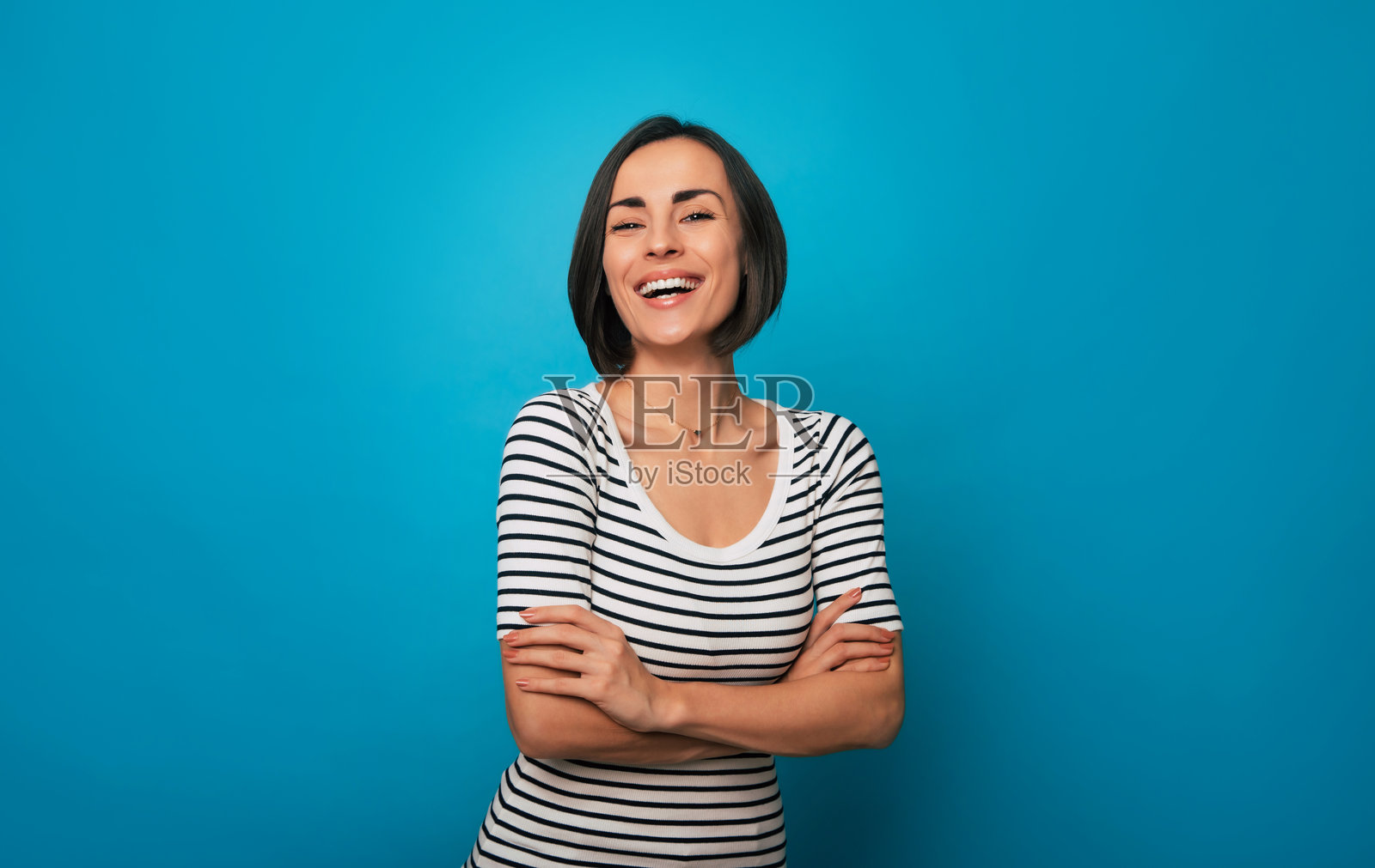 一位短发深色头发的快乐女性穿着条纹衬衫，双臂交叉，站在明亮的蓝色背景前，散发出自信和积极的气息，置身于现代简约的环境中。照片摄影图片