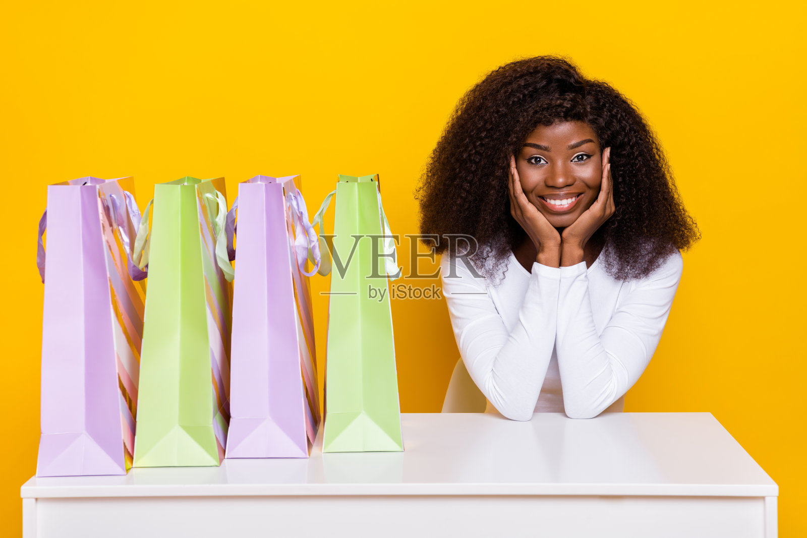 愉快迷人的女士手托着脸颊，桌上放着购物包，背景为黄色。照片摄影图片
