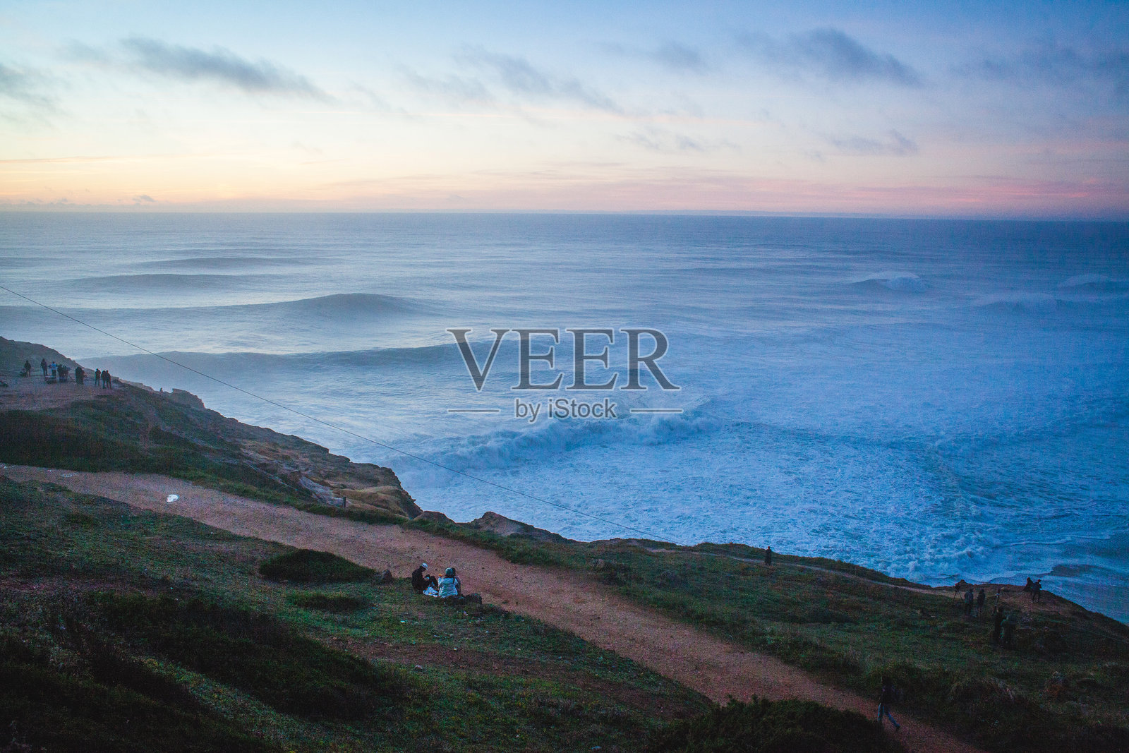 日落时分的海岸景色，波浪拍打着悬崖，人们聚集在一座点亮的建筑附近，营造出宁静而如画的氛围，映衬着崎岖的海岸线。照片摄影图片