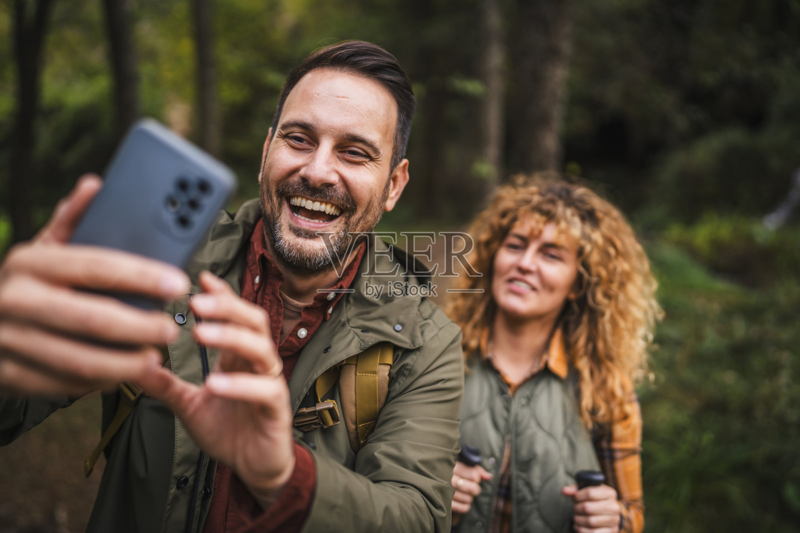 成年男子登山者在拍摄自拍照，他的妻子站在他身后。照片摄影图片