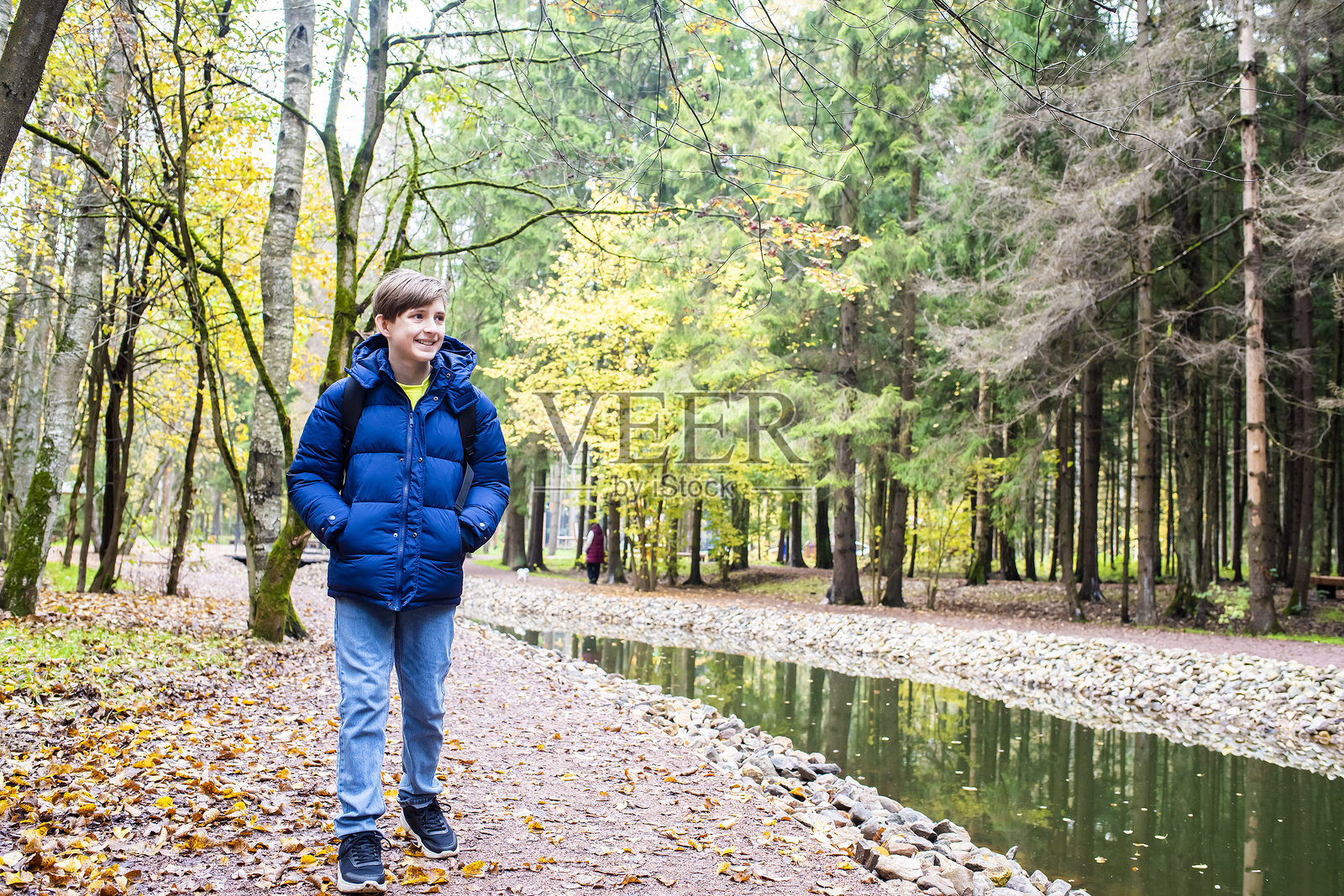 一个穿着蓝色夹克的12岁男孩在秋天的博罗夫斯基公园沿着一系列池塘散步。照片摄影图片