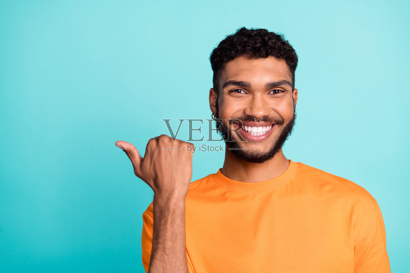 穿着橙色T恤的迷人帅哥在空旷的空间中竖起大拇指，微笑着，背景为青绿色。照片摄影图片