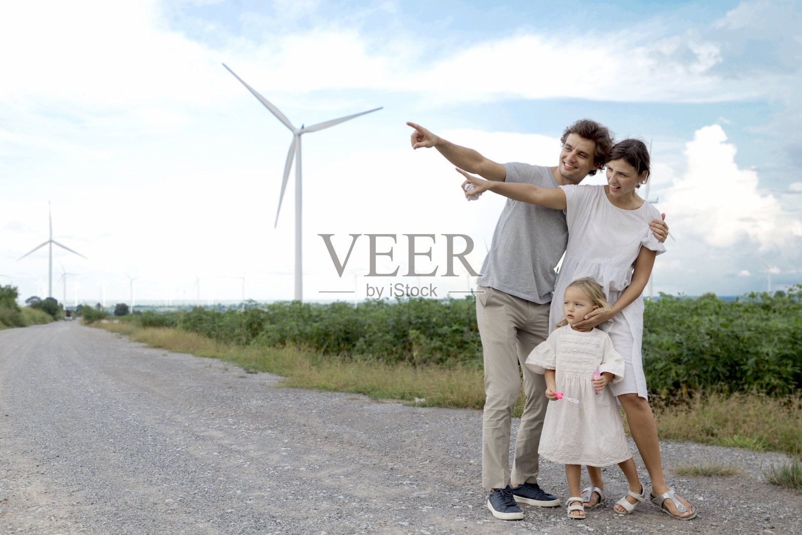快乐的家庭在风力发电场。父亲、母亲和女儿在风车公园享受时光，天空多云。小女孩与父母一起指着远方。生态替代可再生能源。照片摄影图片