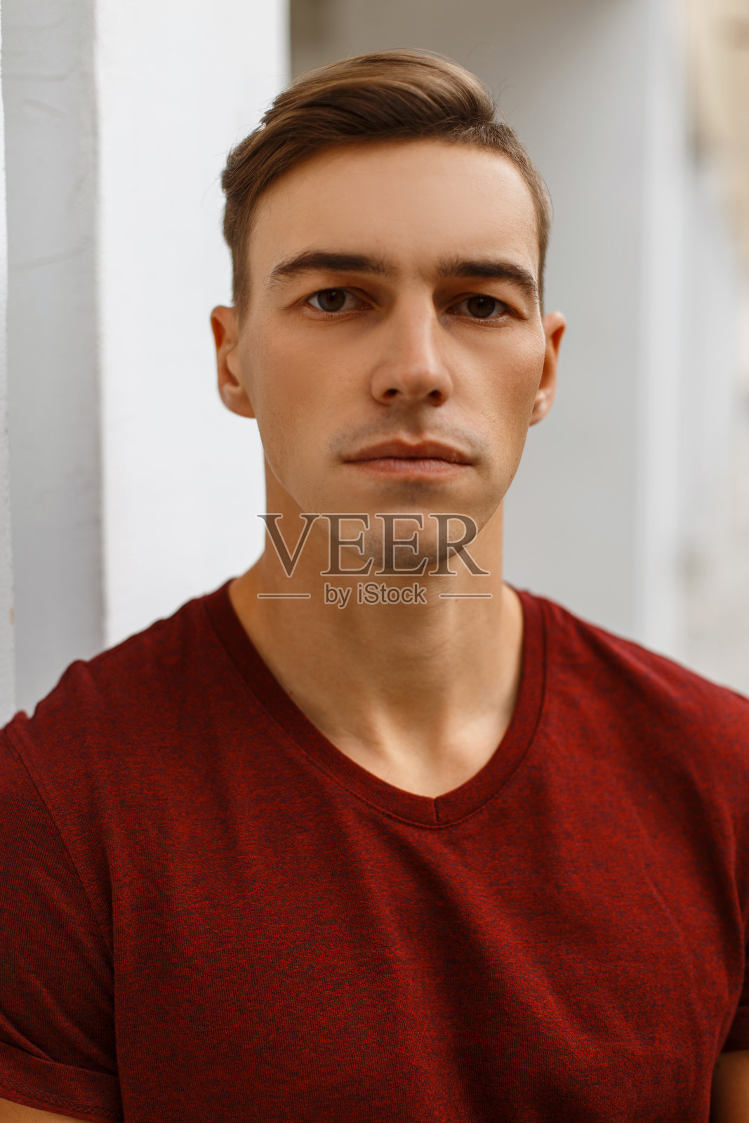 一位严肃的英俊年轻男子，穿着时尚的红色T恤，发型别致，站在一座复古的白色建筑旁。迷人的男士肖像。照片摄影图片