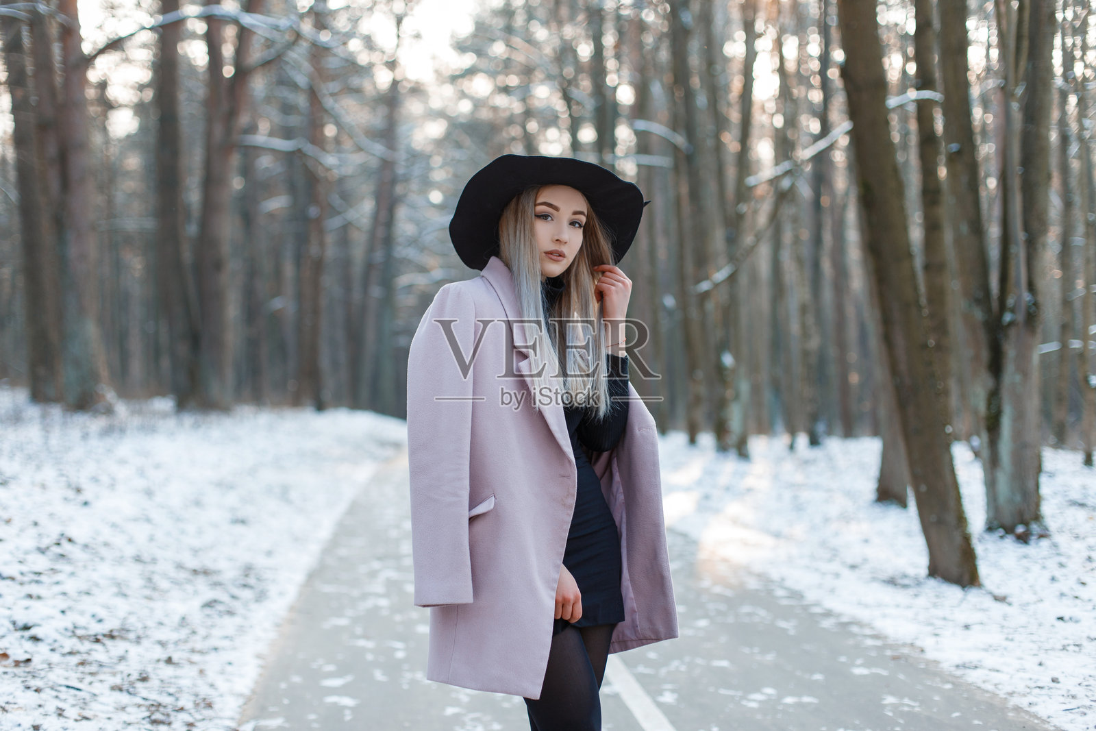 迷人的可爱年轻女性，金发，戴着优雅时尚的帽子，穿着针织高尔夫衫和裙子，身穿时尚的粉色外套，站在雪地公园的小路上，享受温暖的冬日阳光。漂亮的女孩。照片摄影图片