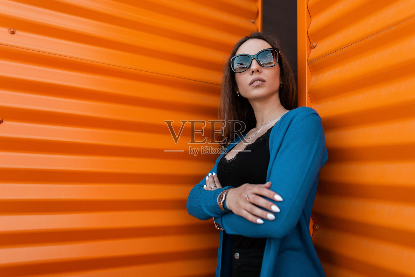 时尚的年轻女性穿着长款针织斗篷，搭配黑色上衣和黑色牛仔裤，戴着复古太阳镜，站在街道上的金属墙旁摆姿势。美丽的女孩在户外放松。照片摄影图片