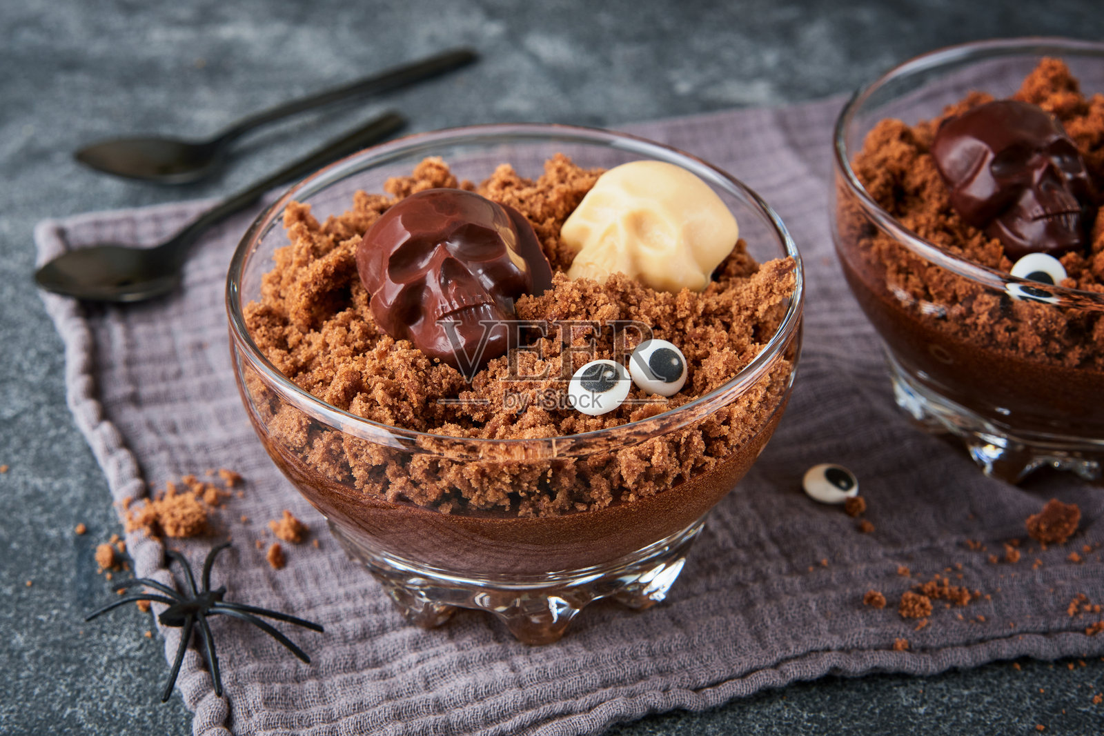 万圣节派对巧克力布丁或意式奶冻，配有布朗尼碎屑和巧克力骷髅，盛放在透明玻璃杯中，背景为深灰色。万圣节派对创意。照片摄影图片