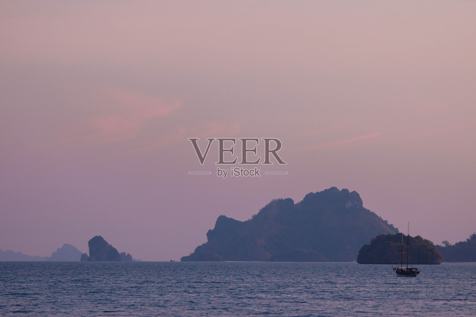 一艘帆船在黄昏时分轻轻滑过平静的水面，远处的岛屿在壮丽的日落映衬下形成剪影，营造出宁静的氛围。照片摄影图片
