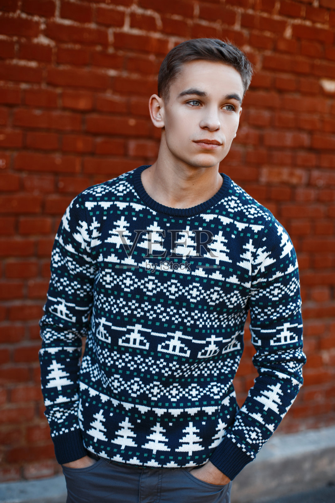 身材帅气、皮肤黝黑的年轻男子，穿着时尚的针织圣诞毛衣和流行裤子，站在街道上，背景是一面砖砌的复古墙壁。美国男孩。照片摄影图片