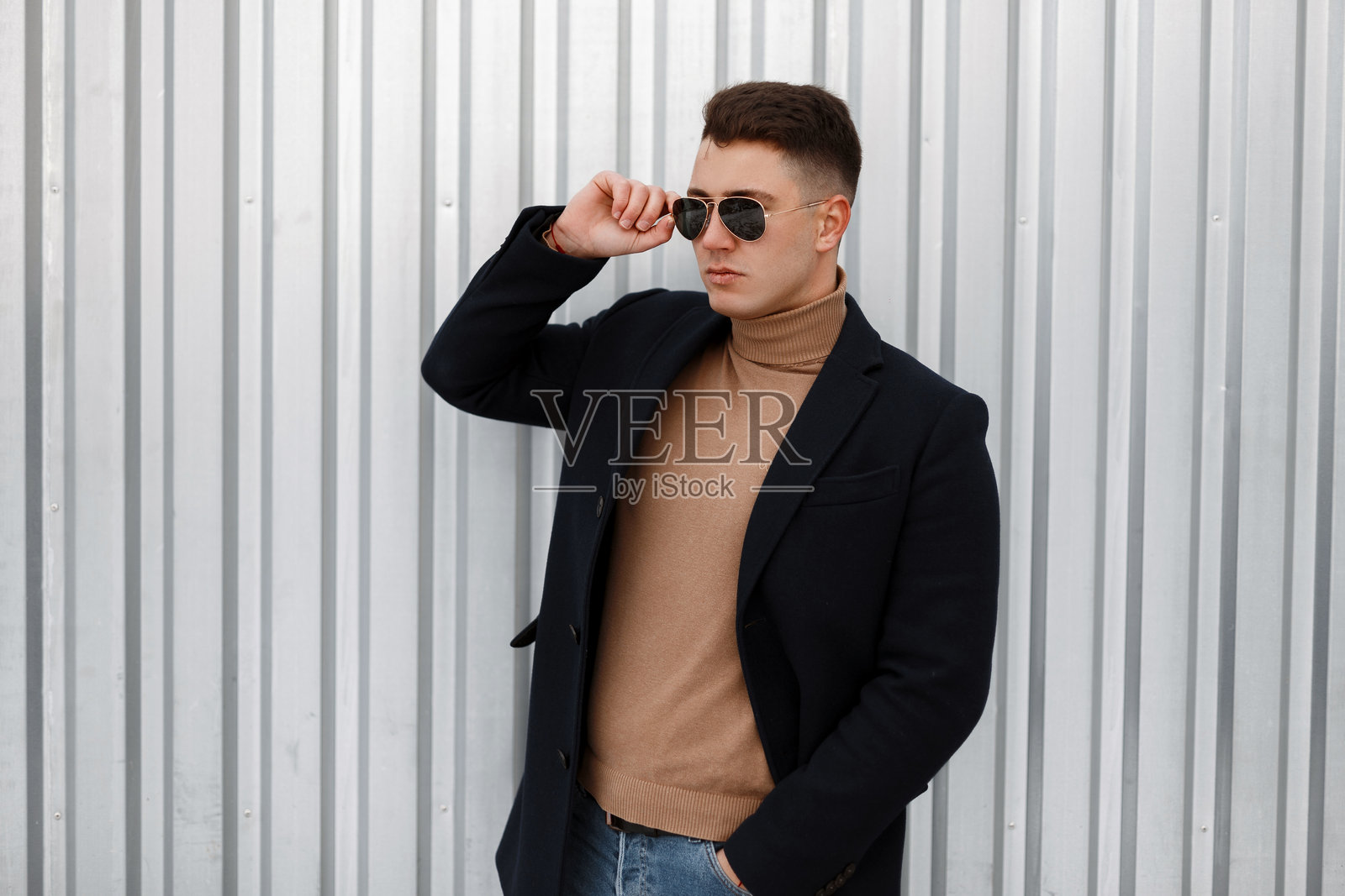 年轻帅气的时尚潮人，穿着秋季时尚外套，搭配米色针织毛衣和时尚太阳镜，牛仔裤，站在户外灰色金属墙旁摆姿势。时尚男士。照片摄影图片