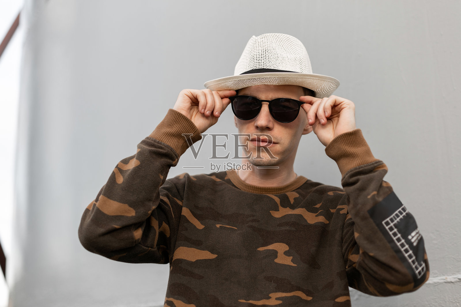 年轻帅气的嬉皮士男士戴着复古帽子，穿着时尚的军装衬衫，站在户外的金属灰色墙壁旁，正在整理时尚的太阳镜。美国现代男模。照片摄影图片