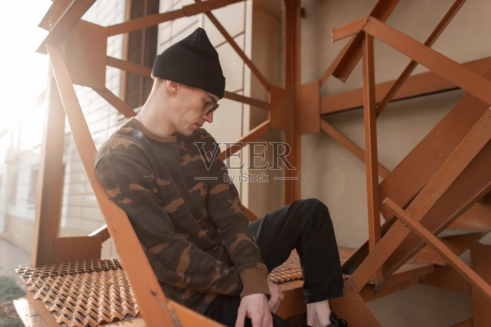 时尚的年轻嬉皮士男士，戴着深色太阳镜，穿着时尚的帽子，身着流行的绿色军装衬衫和牛仔裤，坐在橙色楼梯上放松，享受阳光明媚的日子。美国男孩。街头风格。照片摄影图片