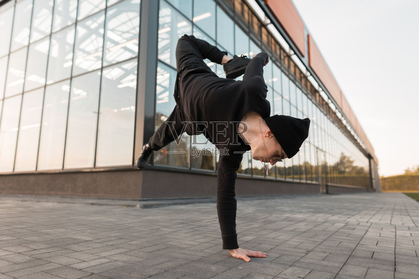 年轻帅气的潮男穿着黑色针织帽、运动鞋、时尚衬衫和牛仔裤，在城市的玻璃建筑旁跳舞。城市男孩在户外做倒立街舞。照片摄影图片