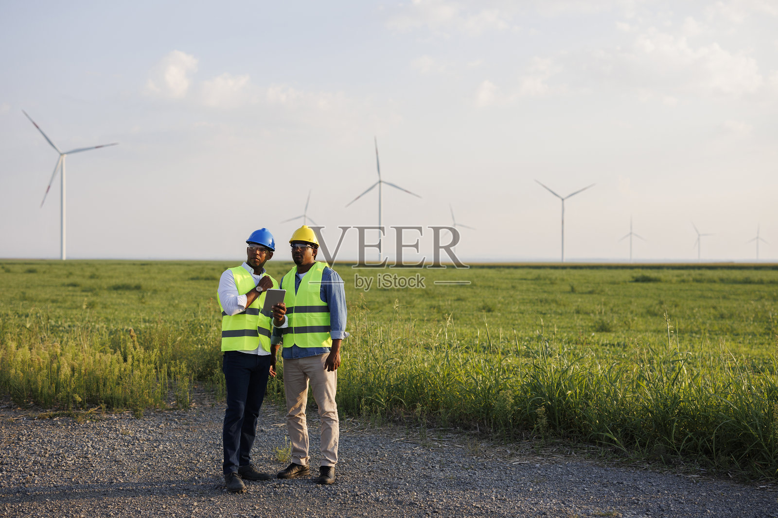 两名年轻工程师穿着制服和安全帽，手持文件在风力发电场进行检查工作。照片摄影图片