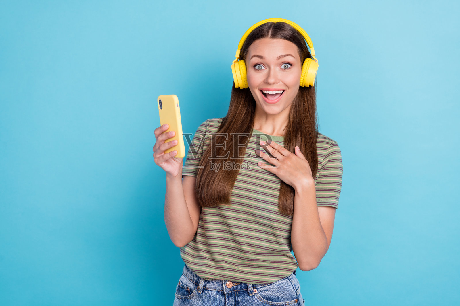 年轻有趣的兴奋女性佩戴新款黄色耳机，手触胸口，手持通知智能手机，背景为蓝色，展示促销信息。照片摄影图片