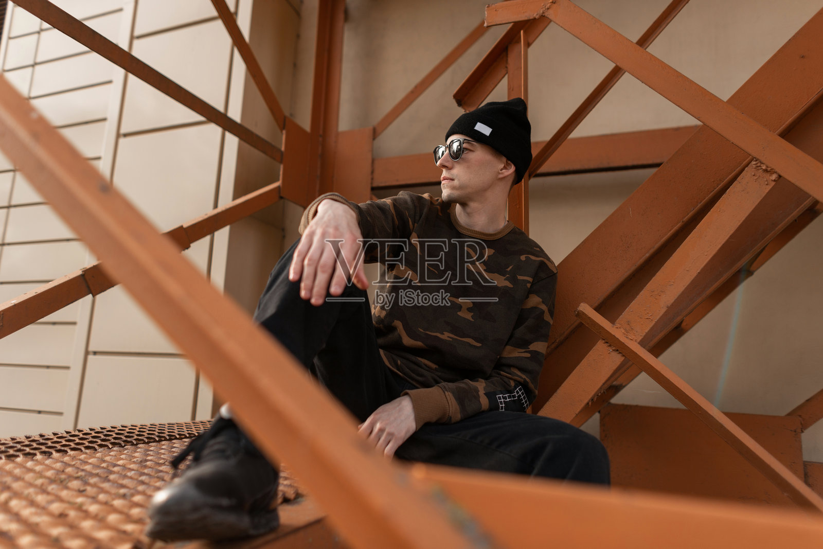 时尚的年轻潮人，戴着黑色帽子，穿着牛仔裤，时尚太阳镜，流行的军装衬衫，搭配皮革运动鞋，坐在街道上的铁制橙色楼梯上休息。城市时尚男士户外服饰。照片摄影图片