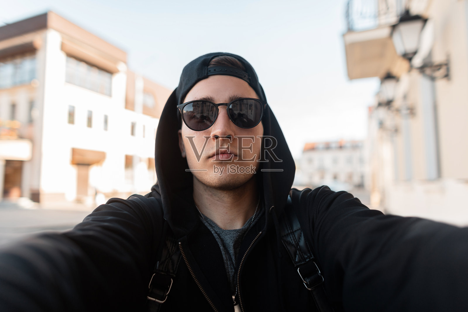 一位严肃帅气的年轻男士，戴着时尚的深色太阳镜和别致的帽子，穿着黑色连帽衫，在阳光明媚的夏日里在城市中自拍。这位友好的美国小伙子走在复古建筑附近。照片摄影图片