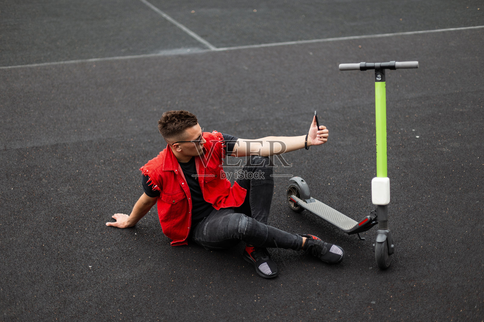 时尚的年轻嬉皮士男士穿着时尚的牛仔服，戴着复古太阳镜，坐在篮球场上用手机自拍。英俊的男孩在户外休息。街头风格。照片摄影图片