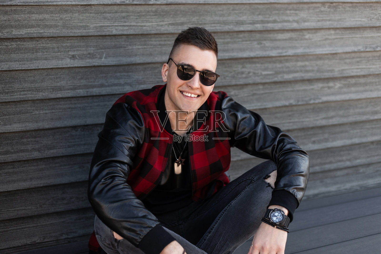 快乐帅气的年轻潮人，穿着时尚的破灰色牛仔裤，搭配红黑格子外套和时尚太阳镜，坐在复古木墙旁，享受周末时光。积极向上的男孩。照片摄影图片
