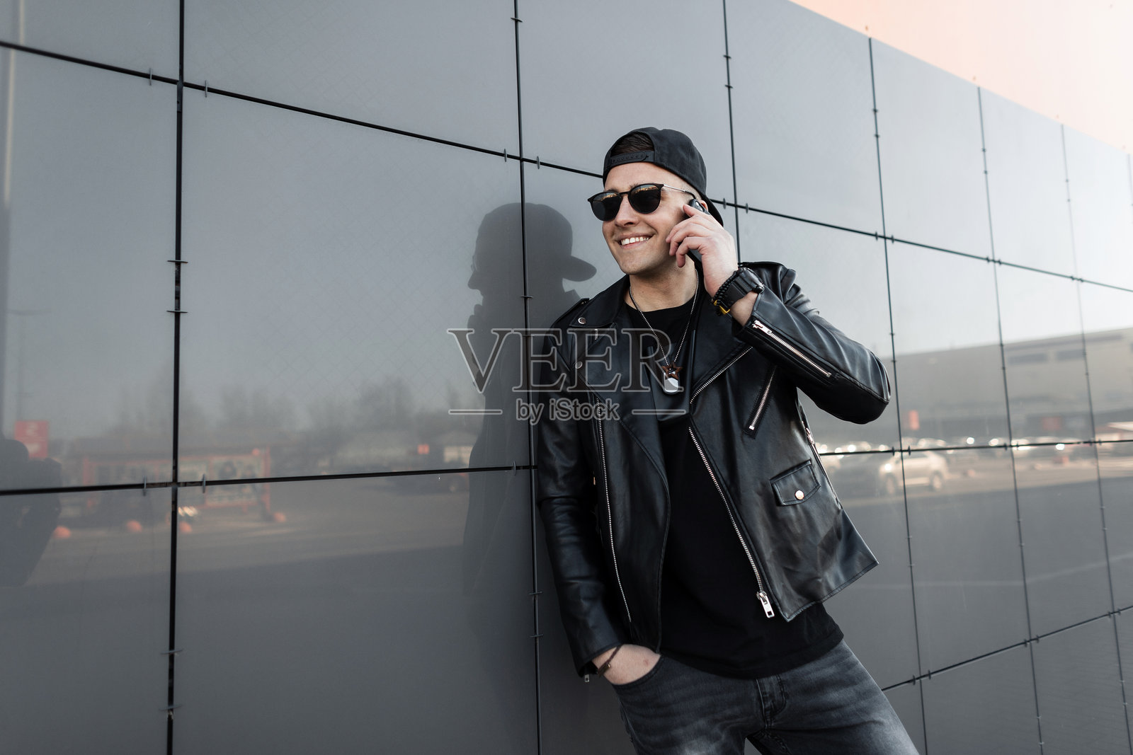 穿着复古皮夹克、T恤和牛仔裤，戴着时尚帽子和深色太阳镜的快乐年轻潮人，微笑着站在现代建筑旁边，正在打电话。帅气的男孩。照片摄影图片